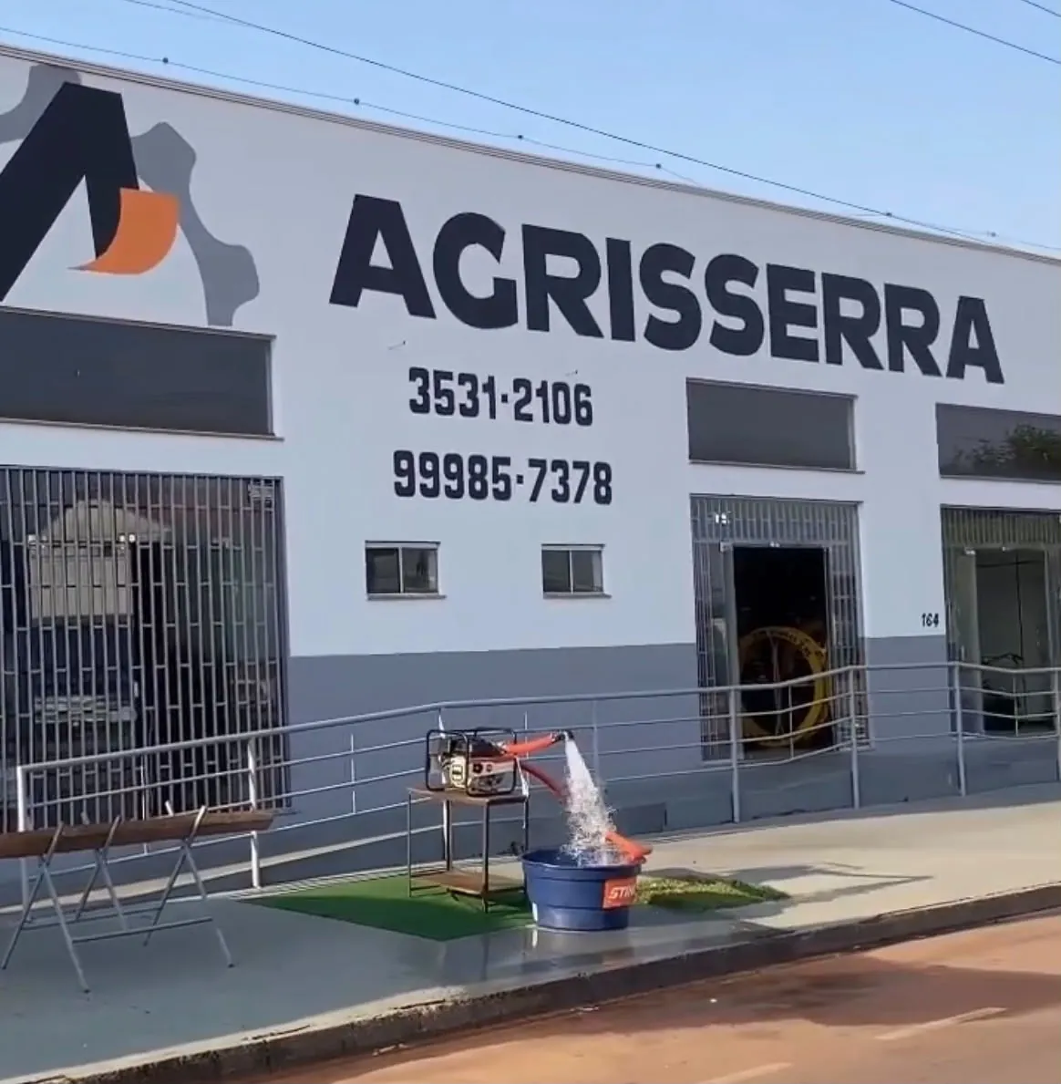 Fachada da Agrisserra na Av. dos Pinheiros: prédio branco com faixa cinza, letreiro, telefones e uma motobomba em demonstração na calçada
